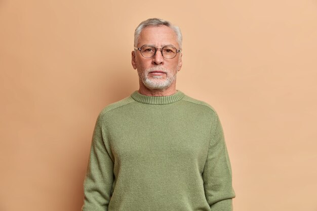 Half length shot of serious bearded man looks unemotional at front with strict expression wears spectacles and jumper has grey hair being confident in something isolated over beige studio wall