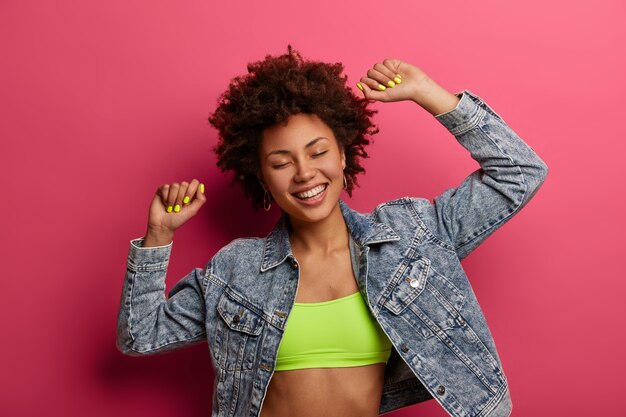 Half length shot of young carefree curly female teenager raises arms, celebrates successfully finished examination session, dances at disco party, makes win gesture, wears top and denim jacket