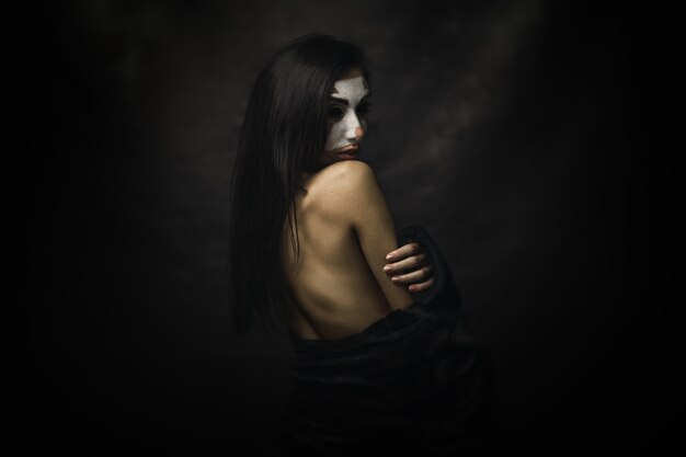 Half-naked female wearing clown makeup on her face standing in front of a black background