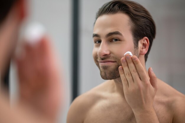 Half-naked man standing near at the mirror and applying shaving foam on his face