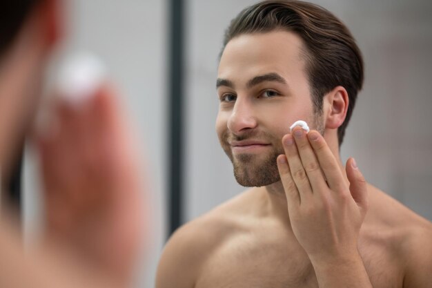 Полуобнаженный мужчина стоит возле зеркала и наносит пену для бритья на лицо