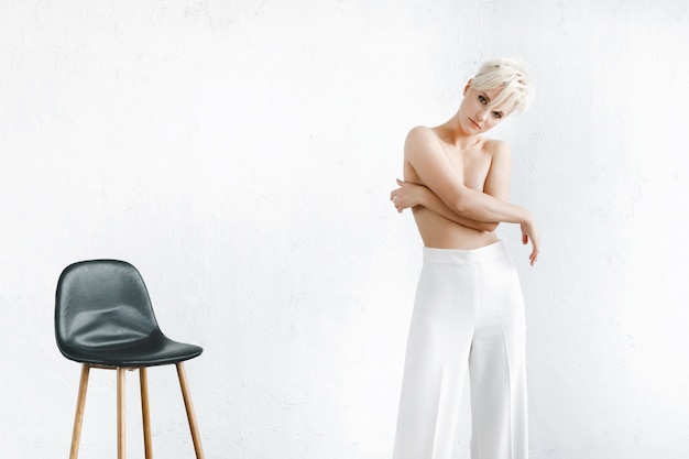 Бесплатное фото Полуголая модель в белых брюках стоит перед белой стеной в студии