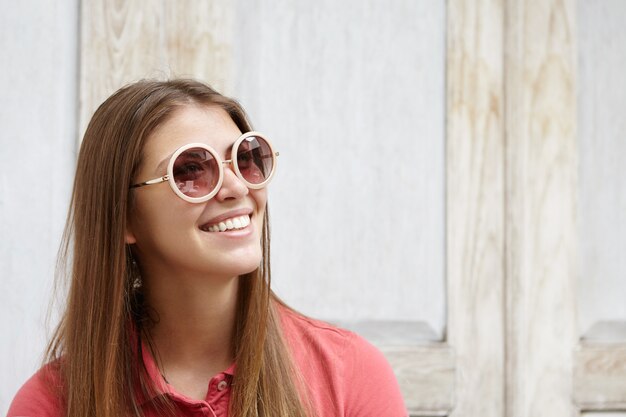 Half profile of attractive fashionable woman in round shades smiling happily showing her white teeth, rejoicing at positive news.