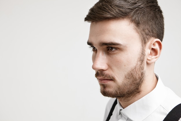 Half profile close up image of self determined focused young Caucasian unshaven businessman in formal clothes posing against white studio wall background with copyspace for your text or content