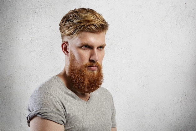 Half-profile   handsome brutal bearded man wearing gray t-shirt with rolled up sleeves looking ahead of him having serious thoughtful face expression. 