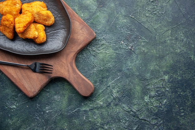 Half shot of chicken nuggets on a black plate and fork on wooden cutting board on the right side on dark surface with free space