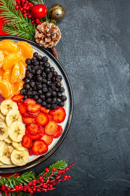 Half shot of collection of fresh fruits on dinner plate decoration accessories fir branches and numbers on a red napkin on a black background