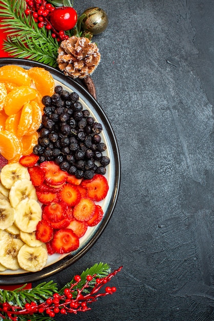 Half shot of collection of fresh fruits on dinner plate decoration accessories fir branches and numbers on a red napkin on a black background