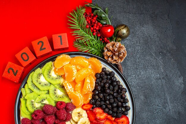 Half shot of collection of fresh fruits on dinner plate decoration accessories fir branches and numbers on a red napkin on a black background