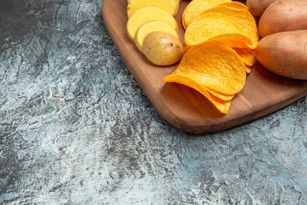 half shot of crispy chips and uncooked potatoes on wooden cutting board on gray table
