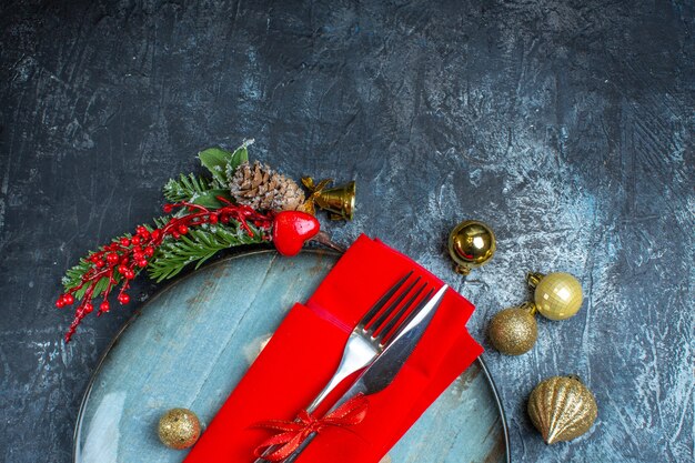 Половина набора столовых приборов с красной лентой на декоративной салфетке на синей тарелке и рождественских аксессуаров на темном фоне