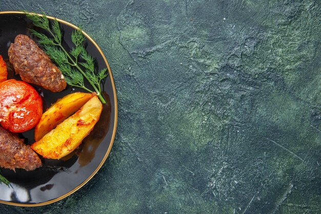 녹색 검정 혼합 색상 배경의 오른쪽에 있는 검정 접시에 감자와 토마토로 구운 맛있는 고기 커틀릿 반샷