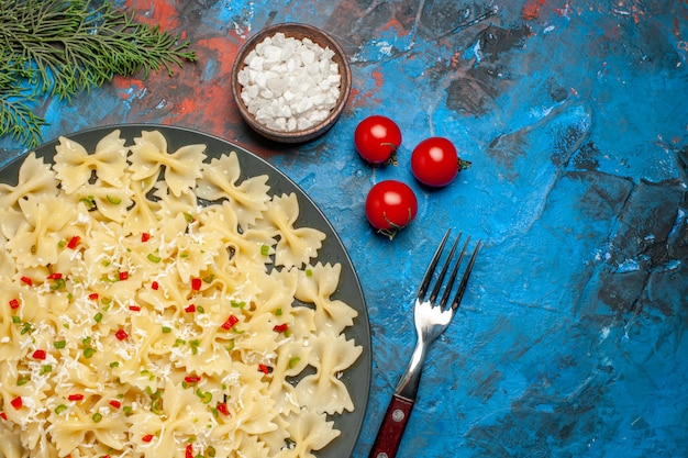 Free photo half shot of farfalle pastas with vegetables tomatoes salt and fork fir branches on blue background