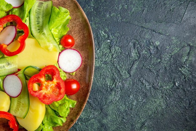 Half shot of fresh peeled cut potatoes with red pepper radishes green tomatoes in a brown plate on the right side on green black mix colors surface