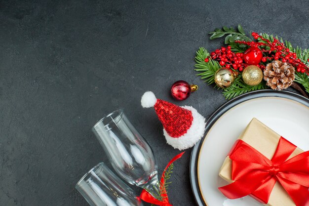 Half shot of gift box on dinner plate christmas tree fir branches conifer cone santa claus hat fallen glass goblets on black background