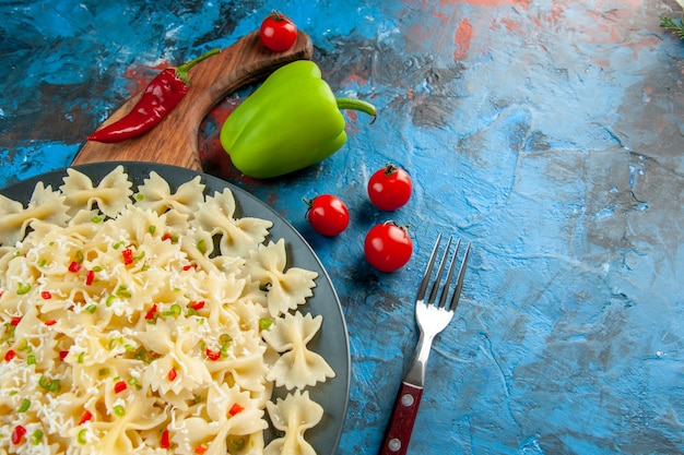 Бесплатное фото Половина выстрела итальянской пасты фарфалле с овощами на черной тарелке на деревянной разделочной доске и вилкой рядом с помидорами перца на синем фоне