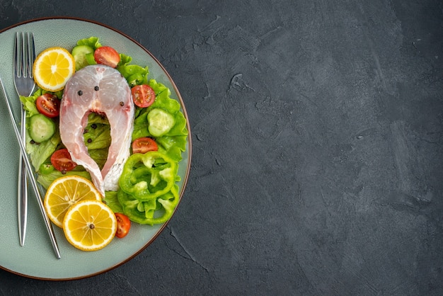 Half shot of raw fish and fresh vegetables lemon slices and cutlery set on a gray plate on the left side on black surface with free space