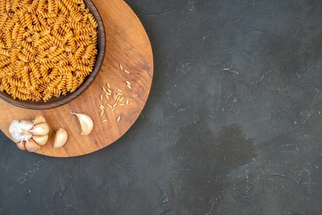 Half shot of raw pastas in a brown bowl garlics rice on wooden board on the right side on black background