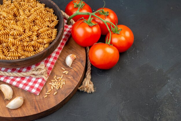 Half shot of raw pastas in a brown bowl on red stripped towel garlics rice on round wooden board tomatoes rope on the right side