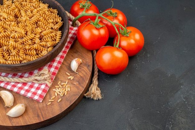 Half shot of raw pastas in a brown bowl on red stripped towel garlics rice on round wooden board tomatoes rope on the right side