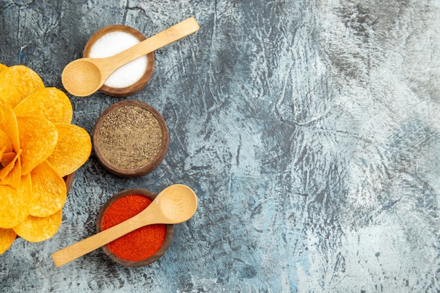 Half shot of tasty potato chips decorated like flower shaped different spices with spoons on them on gray table