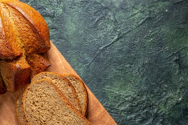 Half shot of whole and cut black bread on brown wooden cutting board on dark colors background