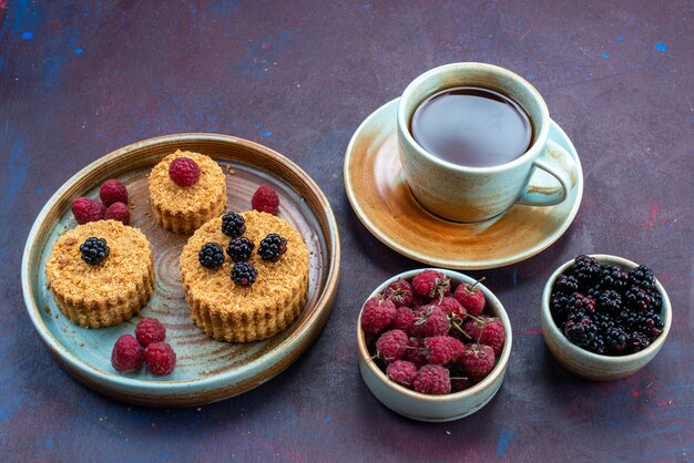 Вид сверху на маленькие сладкие и вкусные пирожные со свежими ягодами и чаем на темной поверхности