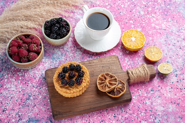 Half-top view raspberries and blackberries inside little pots with cup of tea cake on light-pink background.