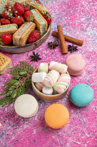 Half-top view of yummy waffle cookies with macarons and fresh red strawberries on pink surface