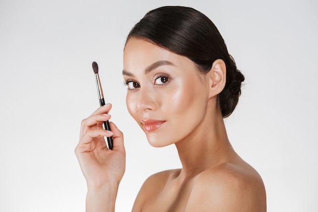 Free photo half-turn picture of pleased woman with fresh skin looking on camera and holding make up brush for eyeshadow, isolated over white wall