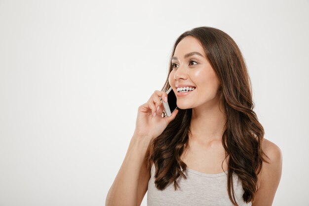 Half-turn portrait of caucasian woman with long brown hair smiling while having pleasant mobile conversation on her smartphone, over white wall