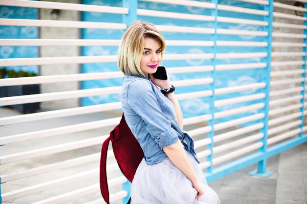 Half-turned beatiful girl with bright pink lips holding smartphone with blue and white stripes on the background.