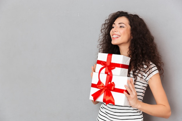 Half turned photo of gorgeous female in striped t shirt holding two gift wrapped boxes with red bows being excited and joyous over grey wall