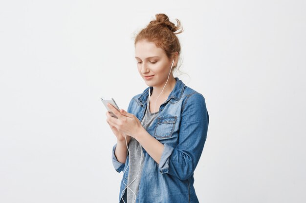 Half-turned portrait of cute young ginger girl with hair combed in bun, texting or browsing via smartphone while standing against gray space