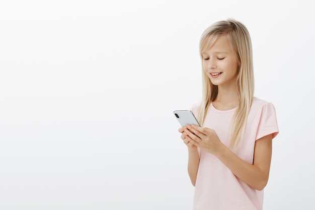 Half-turned profile shot of smart creative female child with blond hair in pink t-shirt, holding smartphone and smiling at screen, playing funny game on device, enjoying spending time over gray wall