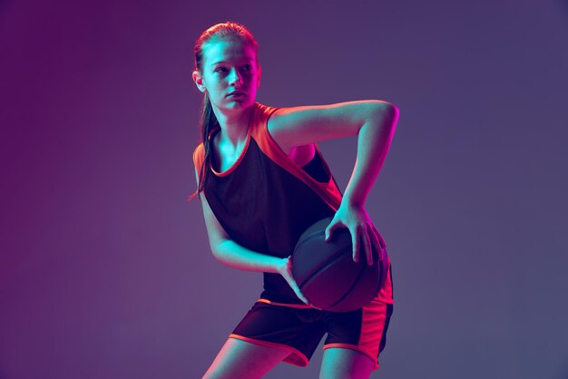 ネオンの紫色の背景の上に分離された若い女の子のバスケットボール選手のトレーニングの半分の長さの肖像画