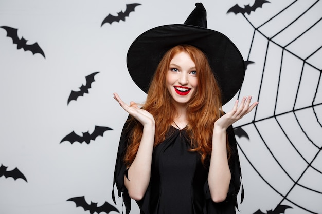 Halloween concept - beautiful caucasian witch surprising with something over grey wall.