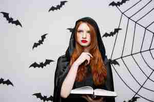 Free photo halloween concept - beautiful dark priest casting spell with magic book on grey wall.