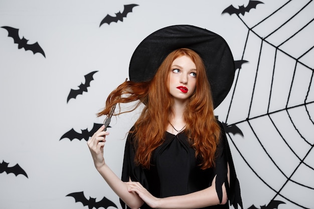 Halloween concept - beautiful witch playing with magic stick on grey wall.