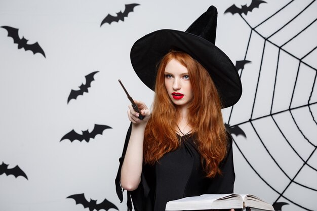 Halloween Concept - Beautiful Witch playing with magic stick on grey wall.