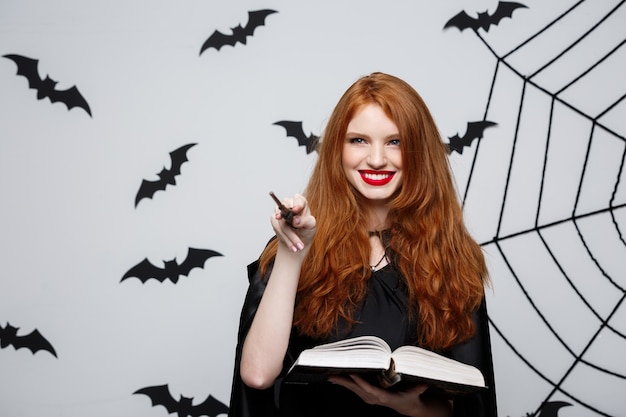 Halloween concept - beautiful witch playing with magic stick and magic book on grey wall.