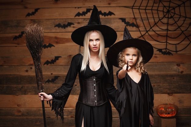 Концепция хэллоуина - жизнерадостная мать и ее дочь в костюмах ведьмы, отмечающих хэллоуин, с изогнутыми тыквами над летучими мышами и паутиной на фоне деревянной студии.