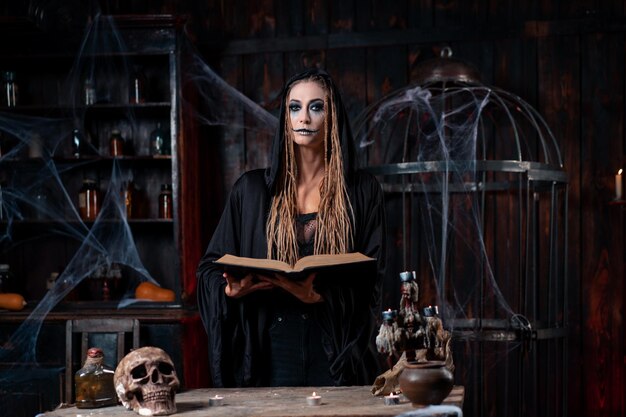 Halloween concept. witch dressed black hood with dreadlocks standing dark dungeon room hold magic book for conjuring curse. female necromancer wizard looking camera with skull, cage, spider web table
