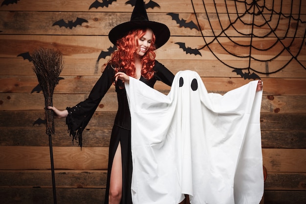 Halloween concept - witch mother and little white ghost doing trick or treat celebrating halloween posing with curved pumpkins over bats and spider web on wooden studio background.