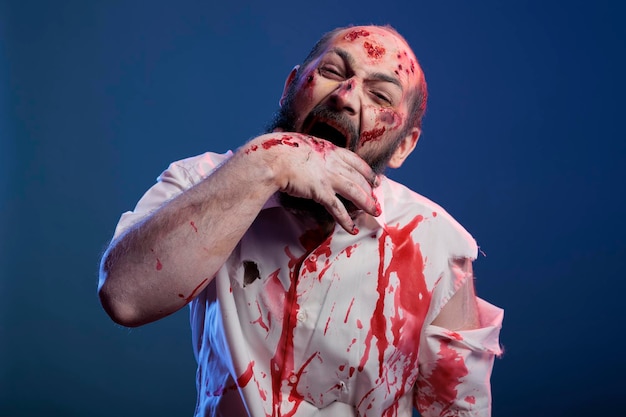 할로윈 잔인한 좀비가 스튜디오에 서서 흉터와 피 묻은 상처로 손을 물어뜯습니다. 치명적인 공포 얼굴과 공격적이고 불길한 눈을 가진 묵시적인 무서운 시체와 뇌를 먹는 악당.