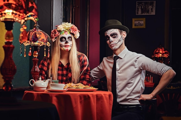 Halloween and Muertos concept. Young attractive couple with undead makeup ordered nachos during dating at a mexican restaurant.