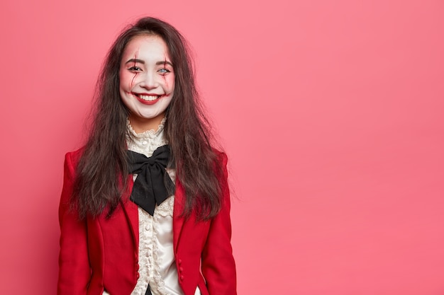 Halloween portrait of cheerful woman with professional makeup being in good mood wears costume and going on masquerade party poses against rosy wall with blank space for your information