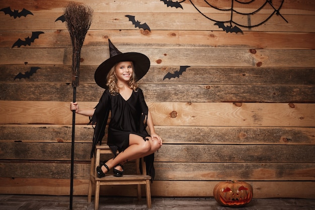 Halloween witch concept - full-length shot of little caucasian witch child posing with magic broomstick over bat and spider web background.
