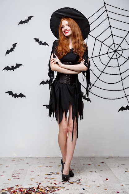 Halloween witch concept  fulllength of happy halloween witch holding posing over dark grey  wall with bat and spider web