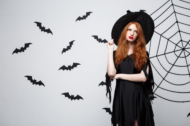 Foto gratuita concetto di strega di halloween strega di halloween che tiene in posa con espressione seria sul muro grigio scuro con pipistrello e ragnatela